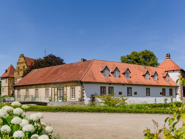 Historisches Gut mit roten Ziegeldächern, umgeben von Grünflächen und blauem Himmel im Hintergrund.