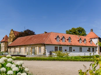 _BBB7003.jpeg Historisches Gut mit roten Ziegeldächern, umgeben von Grünflächen und blauem Himmel im Hintergrund.