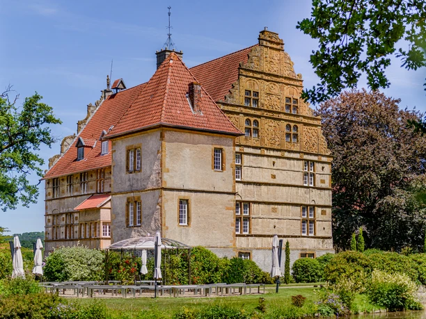 Historisches Schlossgebäude mit markantem roten Dach, umgeben von Garten und Sonnenschirmen.