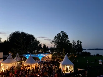 Weinfest-am-Meer-von-oben Ein abendliches Fest am Seeufer mit stimmungsvoller Beleuchtung und zahlreichen Besuchern unter weißen Pagodenzelten.