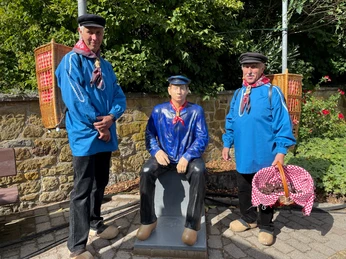 Zwei Männer in traditioneller blauer Kleidung stehen neben einer sitzenden Mann-Statue.