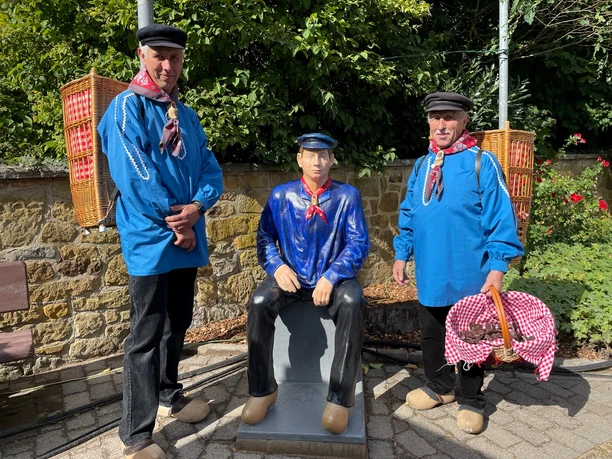 Zwei Männer in traditioneller blauer Kleidung stehen neben einer sitzenden Mann-Statue.
