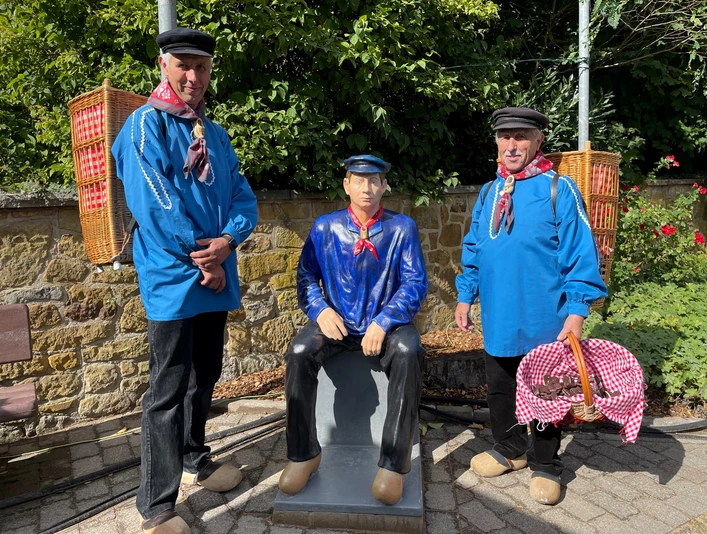 Die Kiepenkerle mit Skulptur.jpg Zwei Männer in traditioneller blauer Kleidung stehen neben einer sitzenden Mann-Statue.