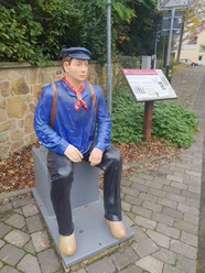 Statue eines Lokführers in traditioneller Uniform, sitzend auf Bank in belebter Fußgängerzone.