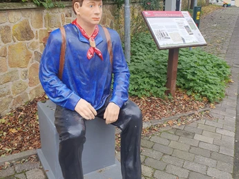 20231106_102208.jpg Statue eines Lokführers in traditioneller Uniform, sitzend auf Bank in belebter Fußgängerzone.
