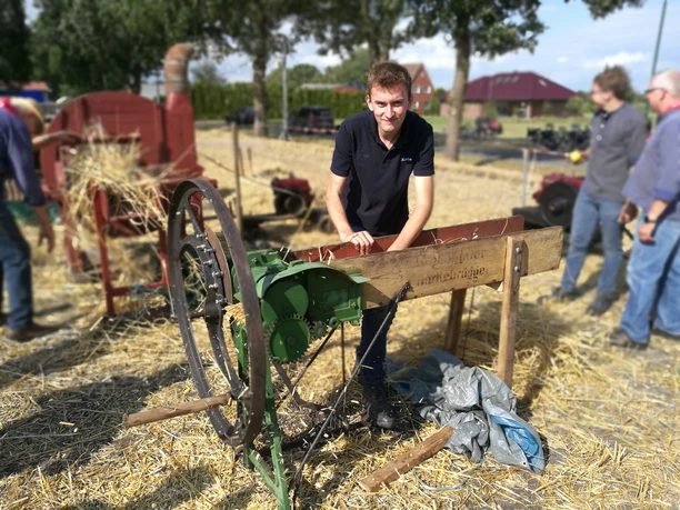 Kornmähen Aperberg Ein junger Mann steht an einer historischen landwirtschaftlichen Maschine zur Strohverarbeitung. Im Hintergrund arbeiten weitere Personen auf einem Strohfeld.