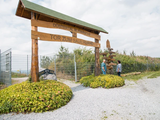 Bad-Wuennenberg_Tor-zum-Sauerland-Ehepaar_16_CC-BY-SA_Bad-Wuennenberg-Touristik-GmbH_Lea-Franke.jpg Ein hölzernes Tor mit der Aufschrift "Tor zum Sauerland" in einer ländlichen Umgebung.