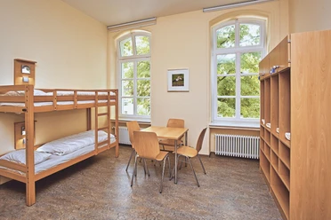 JGH-Petershagen-060_bearbeitet.jpg Ein heller Raum mit Etagenbetten, einem Tisch, Stühlen und großen Fenstern mit Blick ins Grüne.
