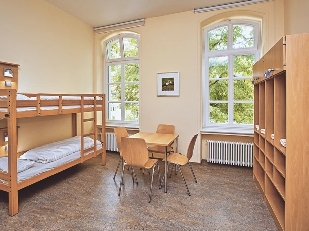 JGH-Petershagen-060_bearbeitet.jpg Ein heller Raum mit Etagenbetten, einem Tisch, Stühlen und großen Fenstern mit Blick ins Grüne.
