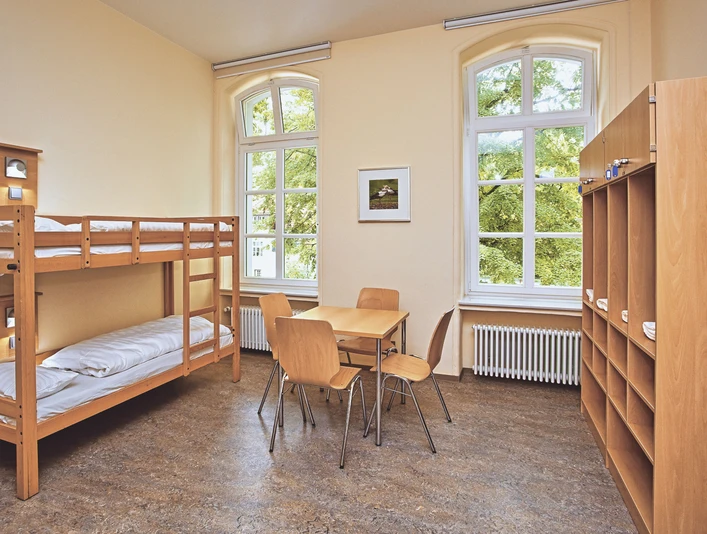 JGH-Petershagen-060_bearbeitet.jpg Ein heller Raum mit Etagenbetten, einem Tisch, Stühlen und großen Fenstern mit Blick ins Grüne.