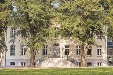 JGH-Petershagen-192_bearbeitet.jpg Historisches Gebäude mit Treppenaufgang, gesäumt von zwei großen Bäumen, im Sonnenschein.