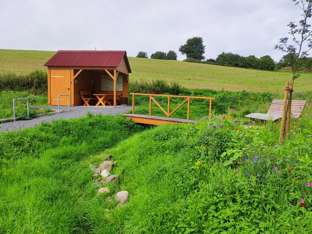Talblick Bärenquelle Holzhütte mit Terrasse und Tischgarnitur, umgeben von Blumenwiese und kleinem Bachlauf.
