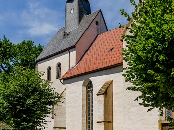 Eine historische Kirche mit spitzem Turm und rotem Ziegeldach, umgeben von grünen Bäumen.