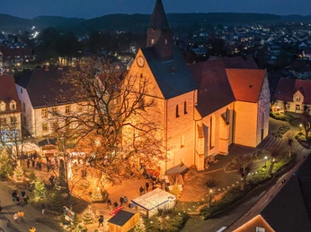 Luftaufnahme eines festlich beleuchteten Weihnachtsmarktes vor einer Kirche in einer abendlichen Szenerie.