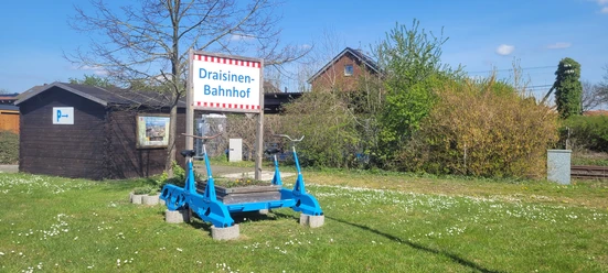 Eine blaue Draisine steht auf einem Rasenstück vor einem Schild mit der Aufschrift „Draisinen-Bahnhof“.