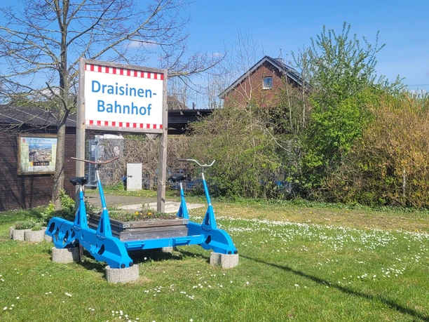 Auenland-Draisinen Eine blaue Draisine steht auf einem Rasenstück vor einem Schild mit der Aufschrift „Draisinen-Bahnhof“.