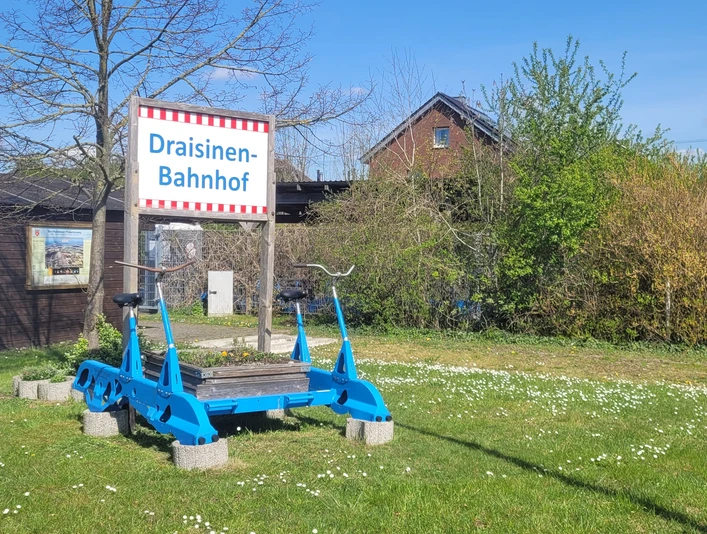 Auenland-Draisinen Eine blaue Draisine steht auf einem Rasenstück vor einem Schild mit der Aufschrift „Draisinen-Bahnhof“.