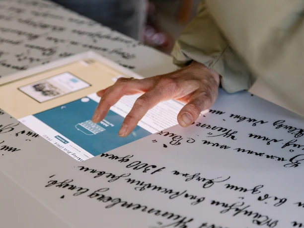 Eine Hand berührt einen Tisch mit altem handgeschriebenem Text, beleuchtet durch ein digitales Display.