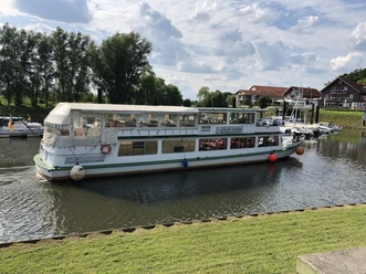 Ein weißes Passagierschiff auf einem ruhigen Fluss neben einem kleinen Hafen mit Gebäuden im Hintergrund.
