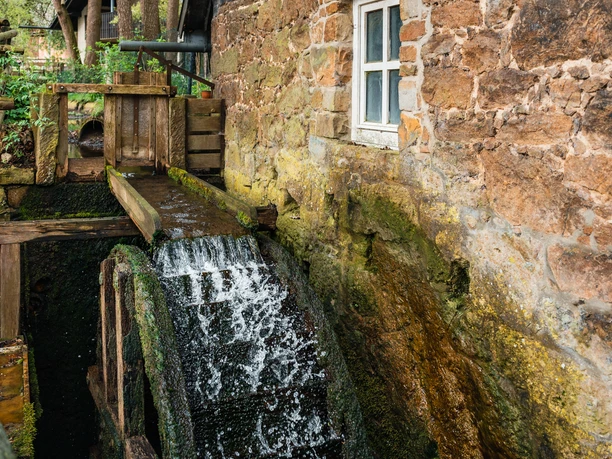 Eine historische Wassermühle mit hölzernem Mühlrad, umgeben von Natursteinen und üppigem Grün.