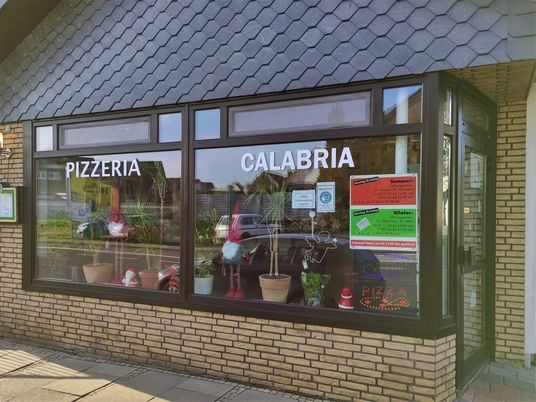 Außenansicht einer Pizzeria mit großen Fenstern, Pflanzen, Weihnachtsdeko und Werbeplakaten.