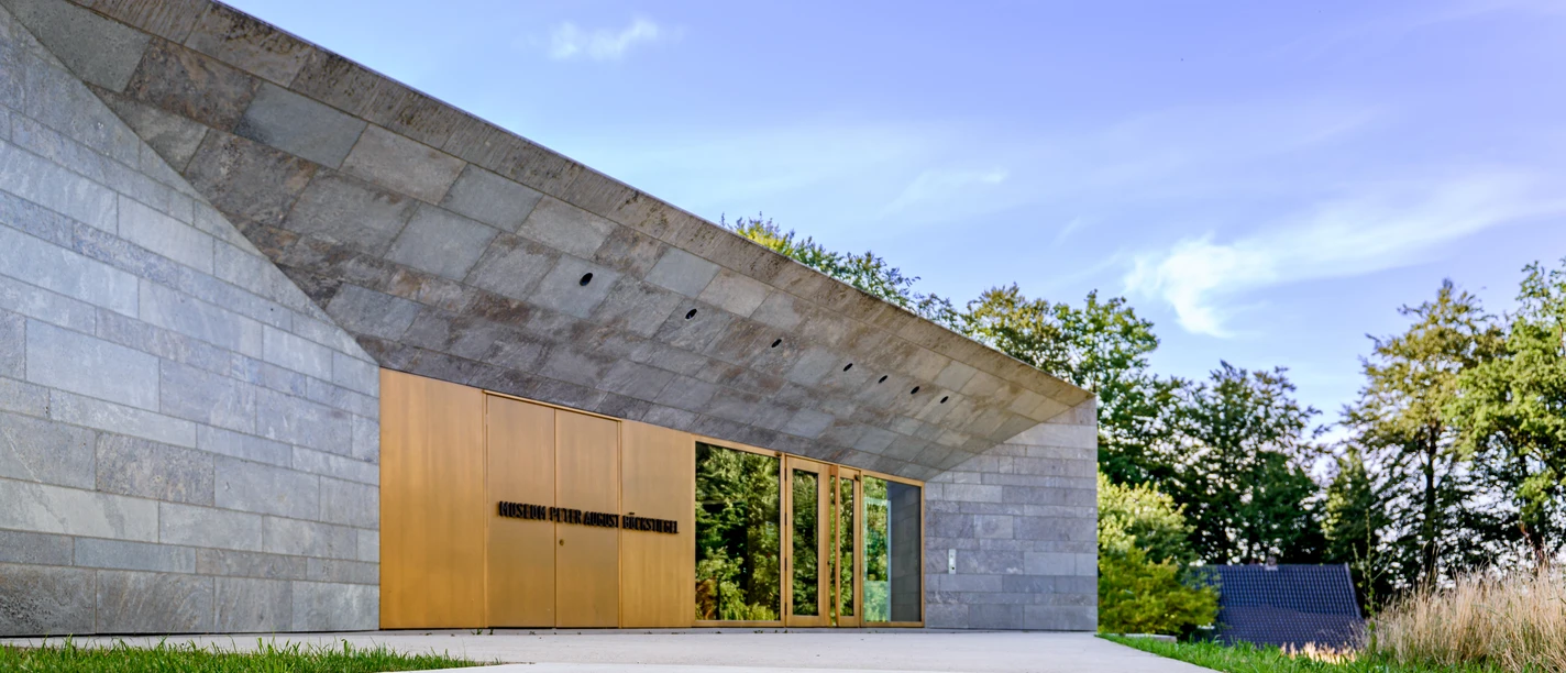 Museum Peter August Böckstiegel Modernes, langes Gebäude mit Stein- und Glaselementen; eingebettet in grüne Landschaft unter blauem Himmel.