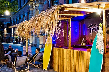 mSa Club Tropicana Sommerfest Sommerfeeling im Club Tropicana: Holzunterstand mit Surfbrettern, im Hintergrund sitzen Personen im Kreis auf Liegestühlen mit Getränken in der Hand