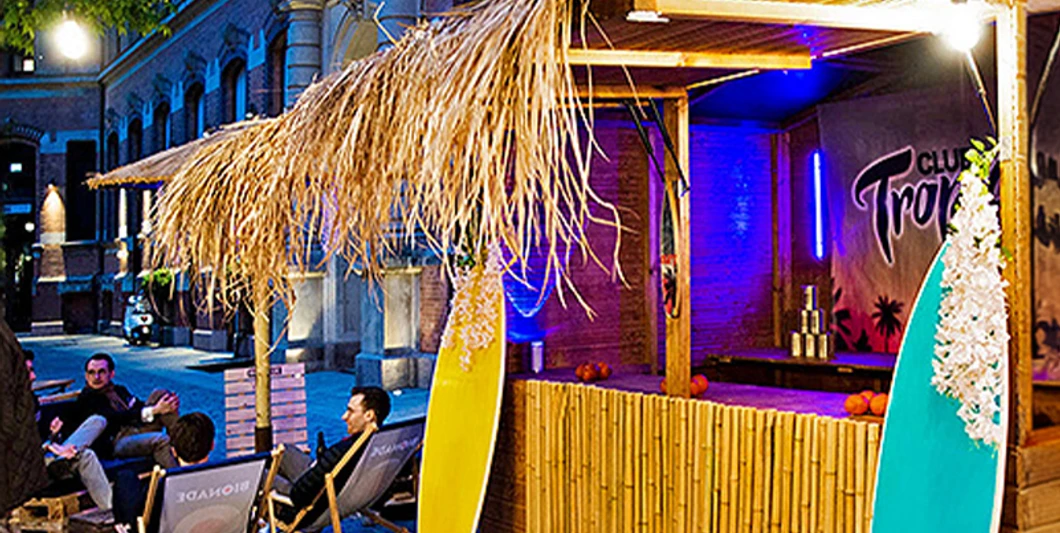 mSa Club Tropicana Sommerfest Sommerfeeling im Club Tropicana: Holzunterstand mit Surfbrettern, im Hintergrund sitzen Personen im Kreis auf Liegestühlen mit Getränken in der HandSummer feeling at Club Tropicana: wooden shelter with surfboards, people sitting in a circle on sun loungers with drinks in their hands in the background