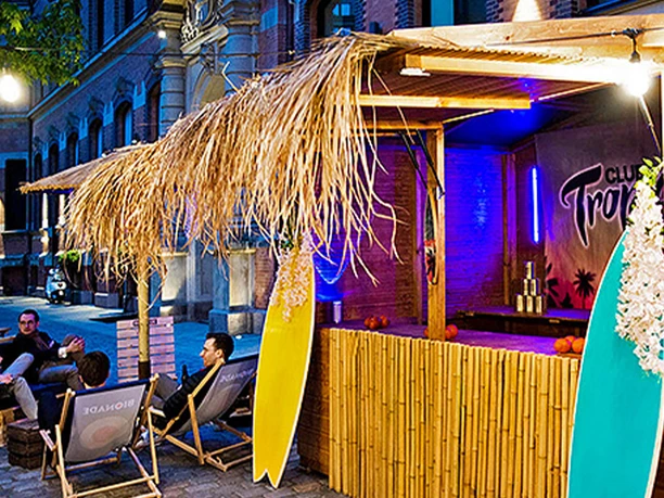 mSa Club Tropicana Sommerfest Sommerfeeling im Club Tropicana: Holzunterstand mit Surfbrettern, im Hintergrund sitzen Personen im Kreis auf Liegestühlen mit Getränken in der Hand