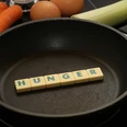 Kochkurs Schwarze Pfanne mit Buchstabensteinen "HUNGER", umgeben von Karotten, Eier und Lauch.