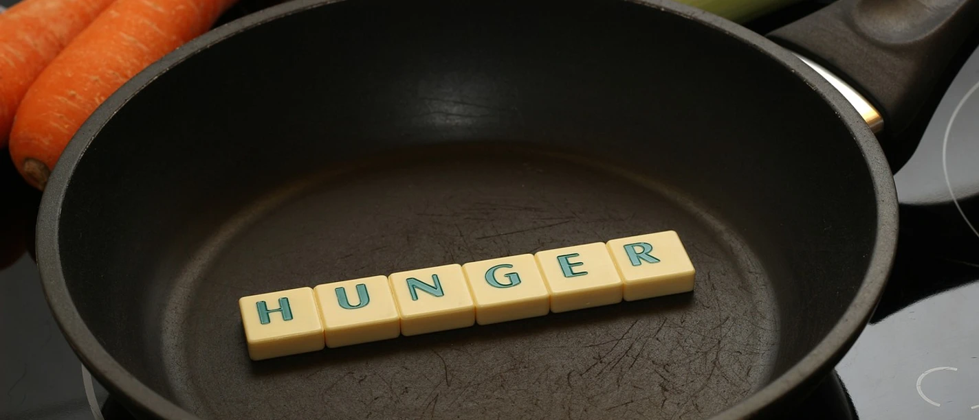 Kochkurs Schwarze Pfanne mit Buchstabensteinen "HUNGER", umgeben von Karotten, Eier und Lauch.