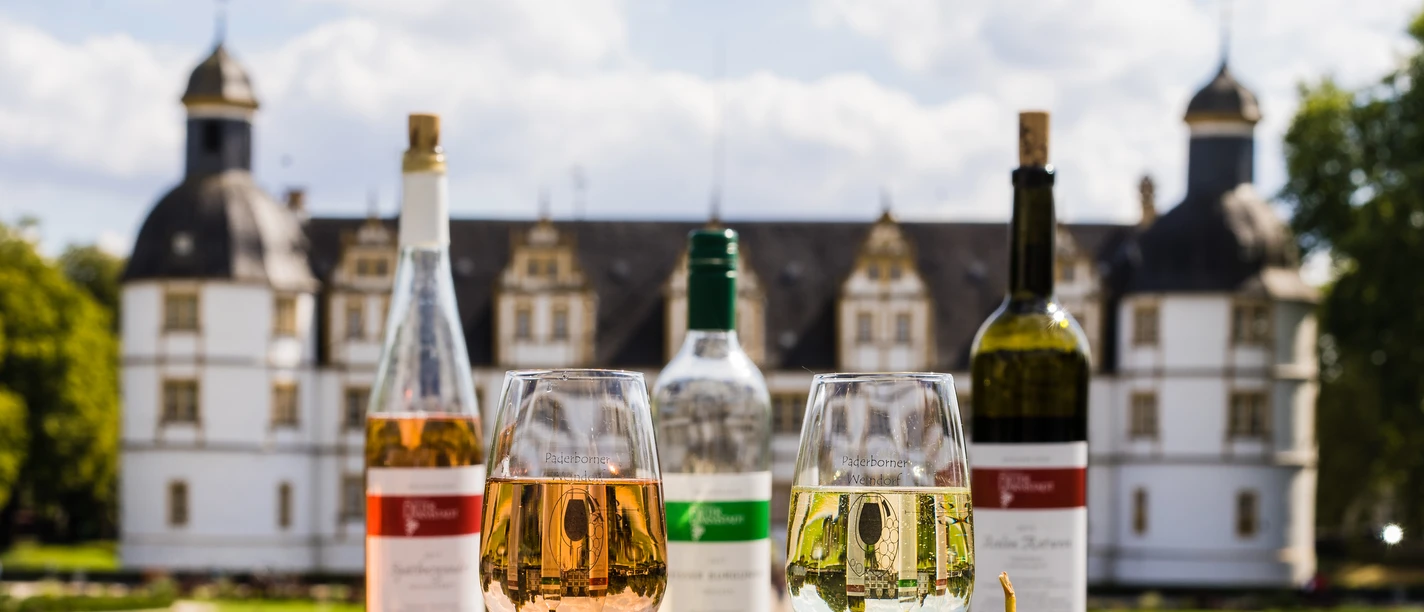 Paderborner Weindorf | Schloss Neuhaus Weingläser und Flaschen vor dem prächtigen Neuhäuser Schloss, umgeben von frischen Trauben.