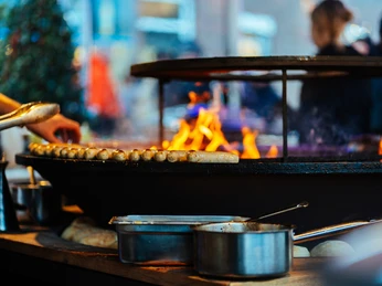 Der Grill in der Gänseliesels Weihnachtslounge Ein großer Grill mit brennendem Holz und darauf befindlichen Bratwürsten, umgeben von Personen.