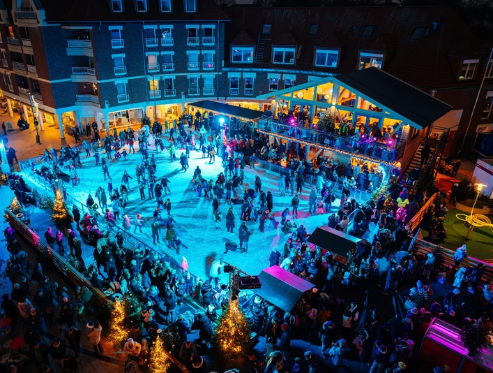 Der Monheimer Sternenzauber mit Eislaufbahn Blick auf eine belebte Eislaufbahn bei Nacht, umgeben von festlich beleuchteten Gebäuden und Bäumen.