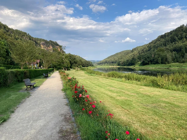 Een pad met bankjes leidt langs een rivier, omzoomd met rode bloemen, omringd door groene heuvels onder een bewolkte hemel.