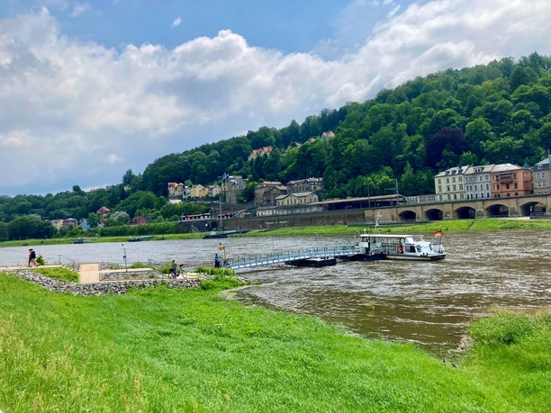 Fähre Königstein-Halbestadt Flusslandschaft mit grünem Ufer, einem Anlegesteg und einem kleinen Boot. Im Hintergrund sind bewaldete Hügel und bunte Gebäude zu sehen.