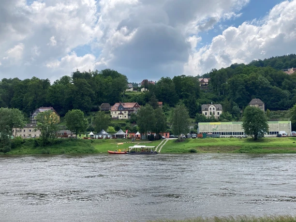 River landscape with a wooded hill in the background, several houses and a tent. The sky is cloudy, the atmosphere is calm.