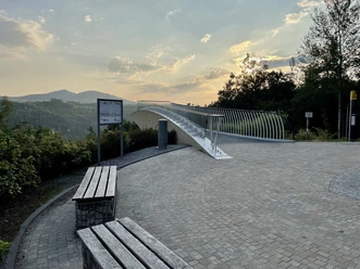 Skywalk Ostrau Moderne Aussichtsplattform mit geschwungenem Geländer, umgeben von Bäumen und Hügeln im Hintergrund bei Sonnenuntergang.