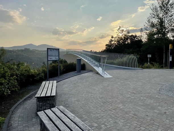 Skywalk Ostrau Modern uitkijkplatform met gebogen leuningen, omringd door bomen en heuvels op de achtergrond bij zonsondergang.
