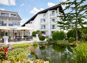 Derichsweiler Hof Ein modernes Hotel mit weißen Fassaden, Balkonen, üppigem Garten und idyllischem Teich davor.