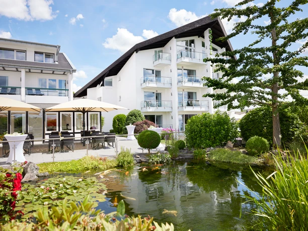Derichsweiler Hof Ein modernes Hotel mit weißen Fassaden, Balkonen, üppigem Garten und idyllischem Teich davor.
