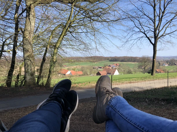 Blick aus der Waldschaukel Zwei Personen entspannen mit Blick auf eine idyllische, von Bäumen umrahmte, grüne Landschaft.