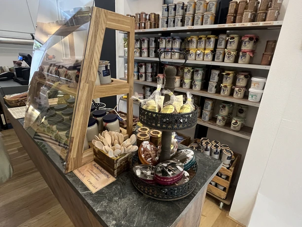 Senfonie 2.jpg Geschäft mit Theke aus Holz und Glas, gefüllt mit verschiedenen Produkten. Regale mit weiteren Produkten im Hintergrund.