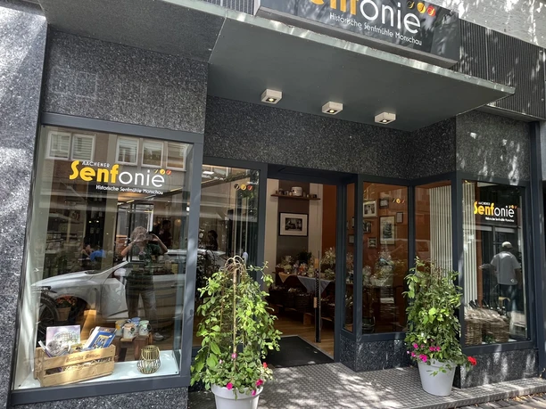 Senfonie 4.jpg Schaufensterfront mit großen Glasflächen, dekoriert mit Pflanzen in eleganten Töpfen. Im Inneren laden helle Räume zum Verweilen ein.