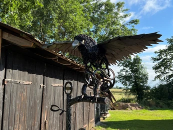 Skulptur Adler Eiserne Adler-Skulptur auf einem Ast vor einer Holzhütte, mit blauen Himmel und Bäumen im Hintergrund.