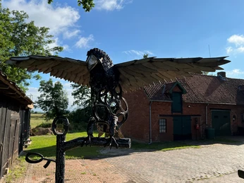 Adlerskulptur mit ausgebreiteten Flügeln, aus Metall gefertigt, vor einem ländlichen Backsteingebäude.