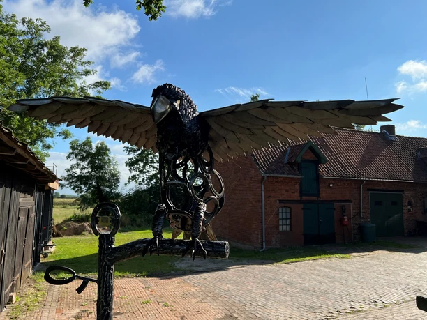 Adlerskulptur mit ausgebreiteten Flügeln, aus Metall gefertigt, vor einem ländlichen Backsteingebäude.