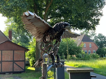 Adlerskulptur aus Metall mit ausgebreiteten Flügeln vor ländlicher Backsteinarchitektur und Bäumen.