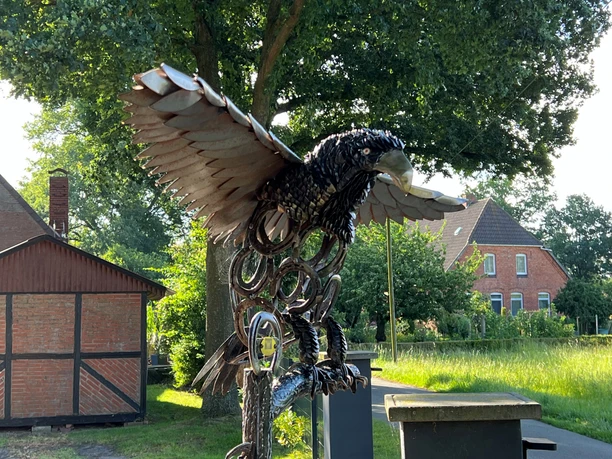 Adlerskulptur aus Metall mit ausgebreiteten Flügeln vor ländlicher Backsteinarchitektur und Bäumen.