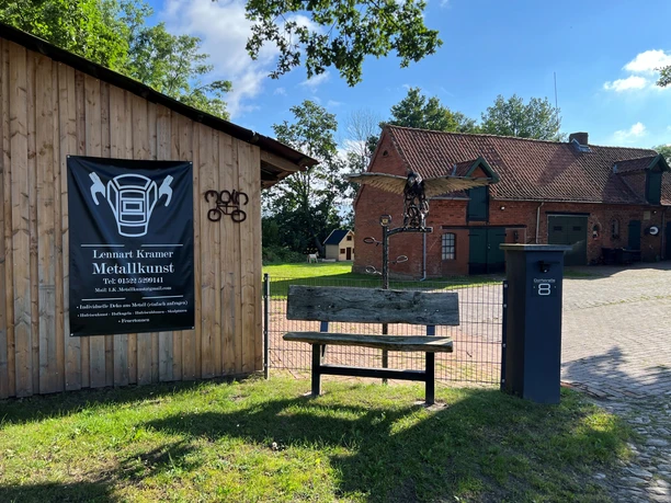 Metallene Skulptur eines Adlers auf einem Holzpfosten vor einem Gebäude, umgeben von grünem Gelände.